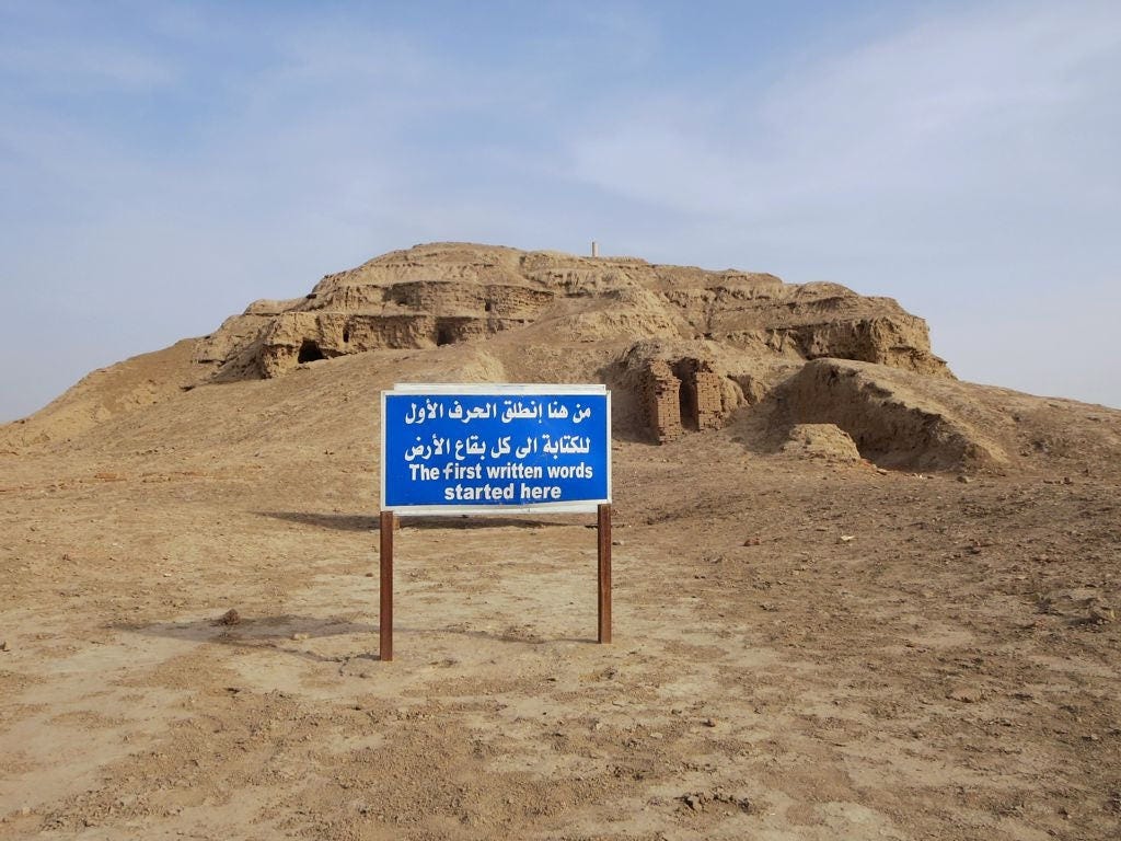 A large platform of mud bricks, eroded and slumping through time, with sloping sides and what look like they were staircases ascending its sides. A blue sign with white lettering in Arabic and English is planted in the foreground. It reads: The first written words started here A large platform of mud bricks, eroded and slumping through time, with sloping sides and what look like they were staircases ascending its sides. A blue sign with white lettering in Arabic and English is planted in the foreground. It reads: The first written words started here