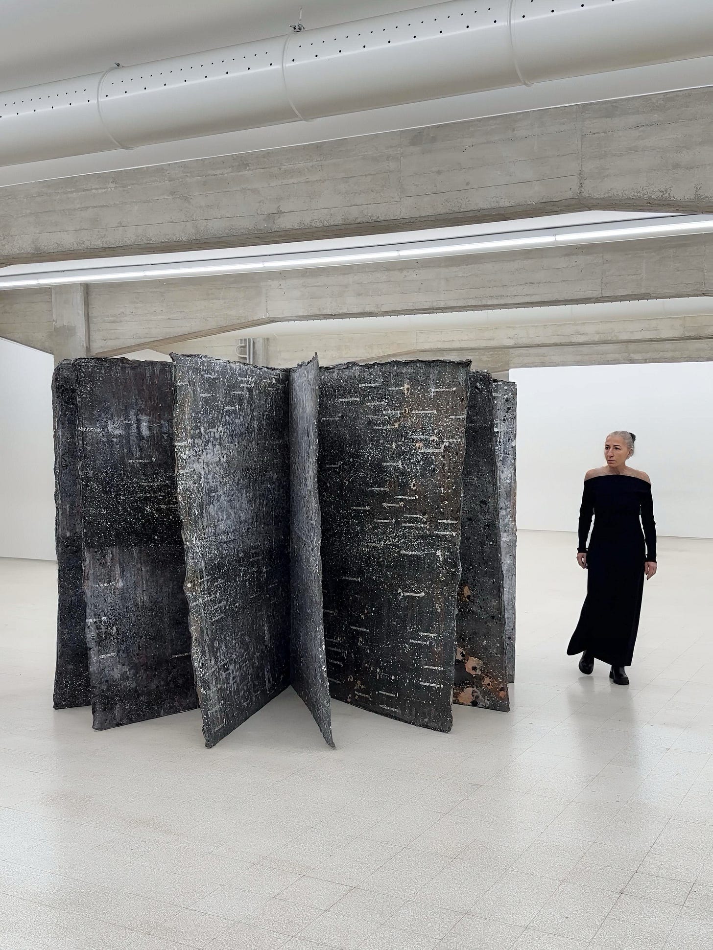 Roberta Bertozzi standing beside a monumental lead book installation by Anselm Kiefer, in a white museum space. The image emphasizes scale, material weight, and the impossibility of reading as a physical experience.