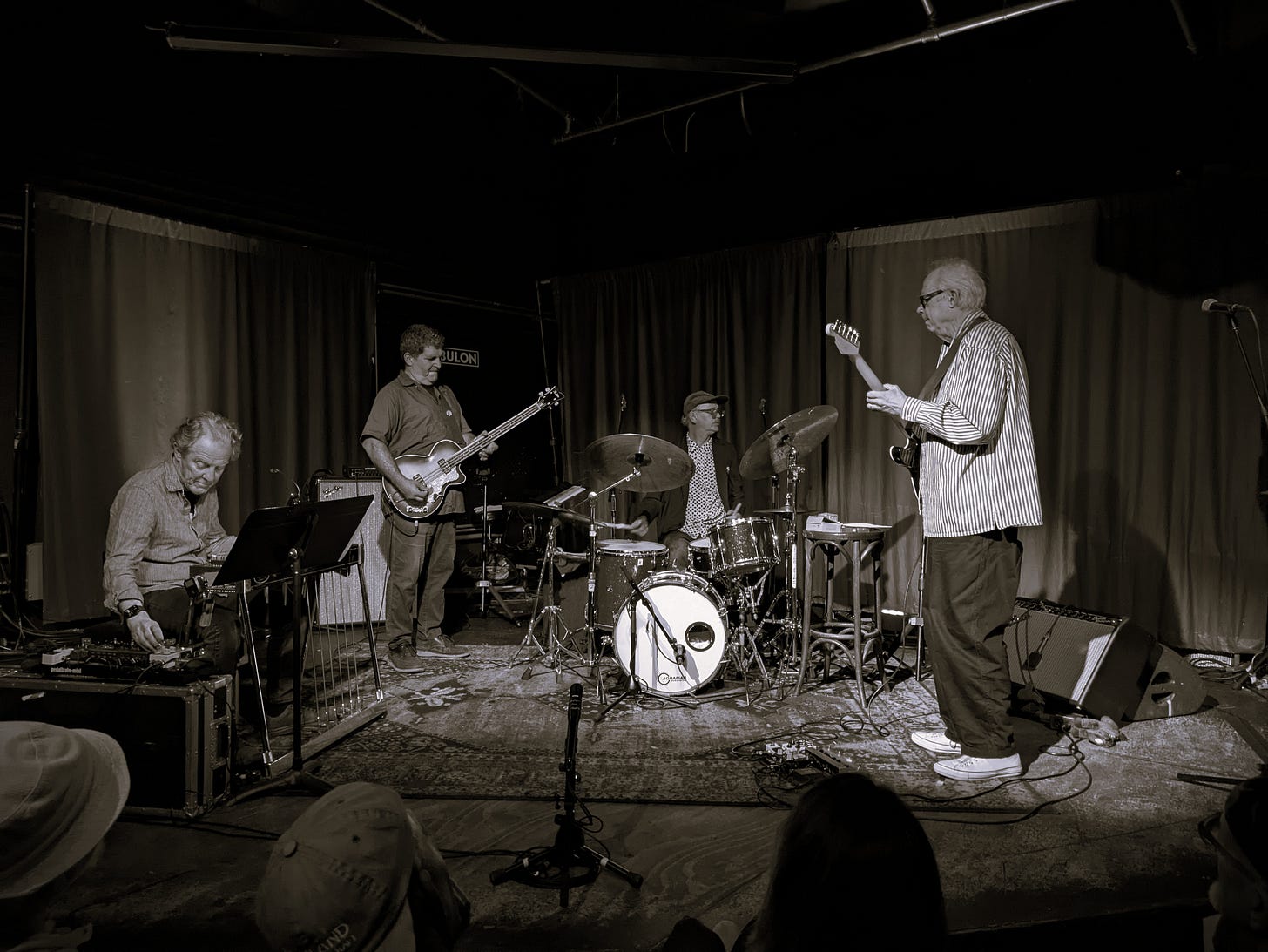 Black and white photo of Bill Frisell playing the electric guitar with his band members on a small stage.
