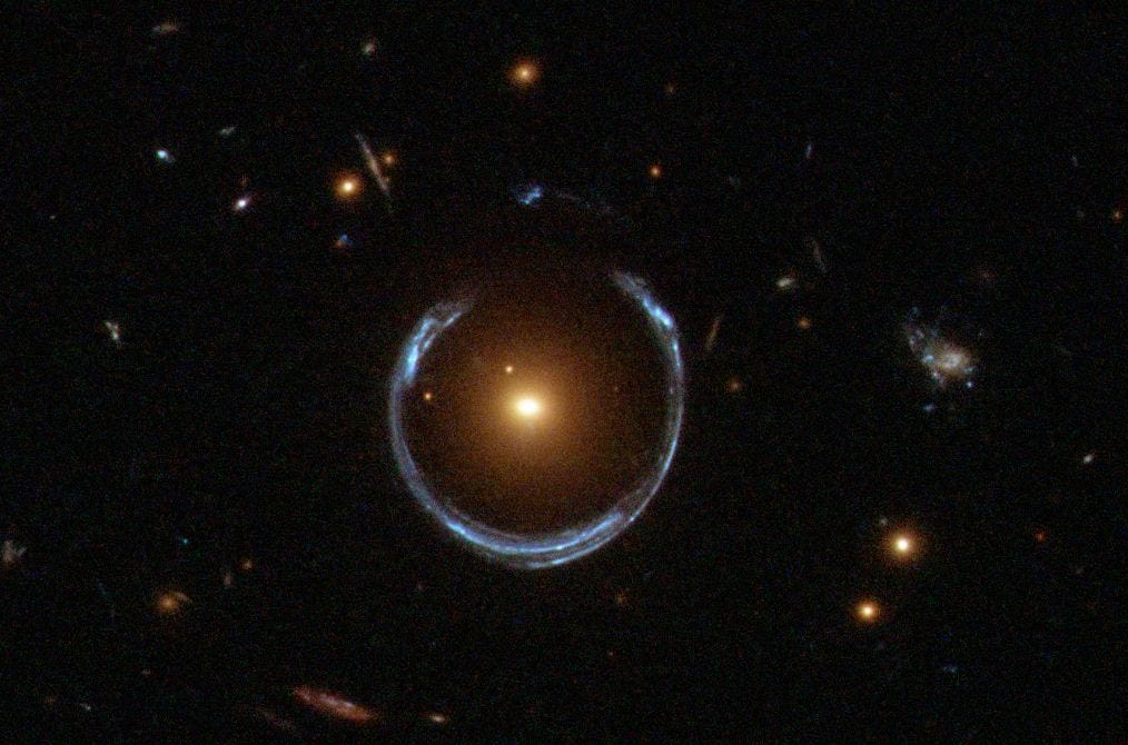 A Hubble photograph of a blue horseshoe-shaped arc of light wrapped almost entirely around a compact red galaxy. A Hubble photograph of a blue horseshoe-shaped arc of light wrapped almost entirely around a compact red galaxy.