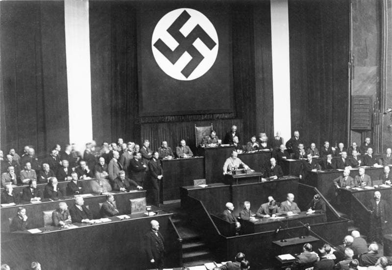 Adolf Hitler speaks from the rostrum of a crowded parliamentary chamber in the Kroll Opera House, with a huge swastika banner hanging above the presiding officers.