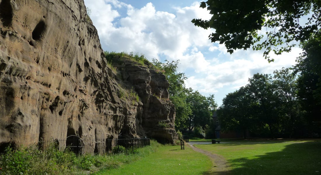 A wall of caves with a grass lawn next to it, and a blue sky with clouds above it