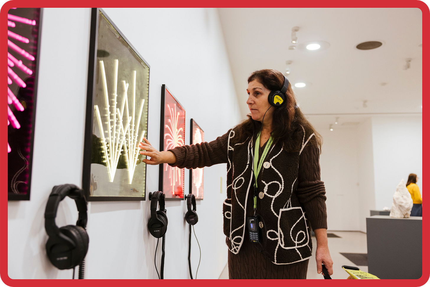 A photo of a blind woman wearing yellow-black headphones while reaching out her hand to touch a bright neon artwork on the gallery wall. In her other hand, she is holding a dog lead; her guide dog is just out of shot. A photo of a blind woman wearing yellow-black headphones while reaching out her hand to touch a bright neon artwork on the gallery wall. In her other hand, she is holding a dog lead; her guide dog is just out of shot.