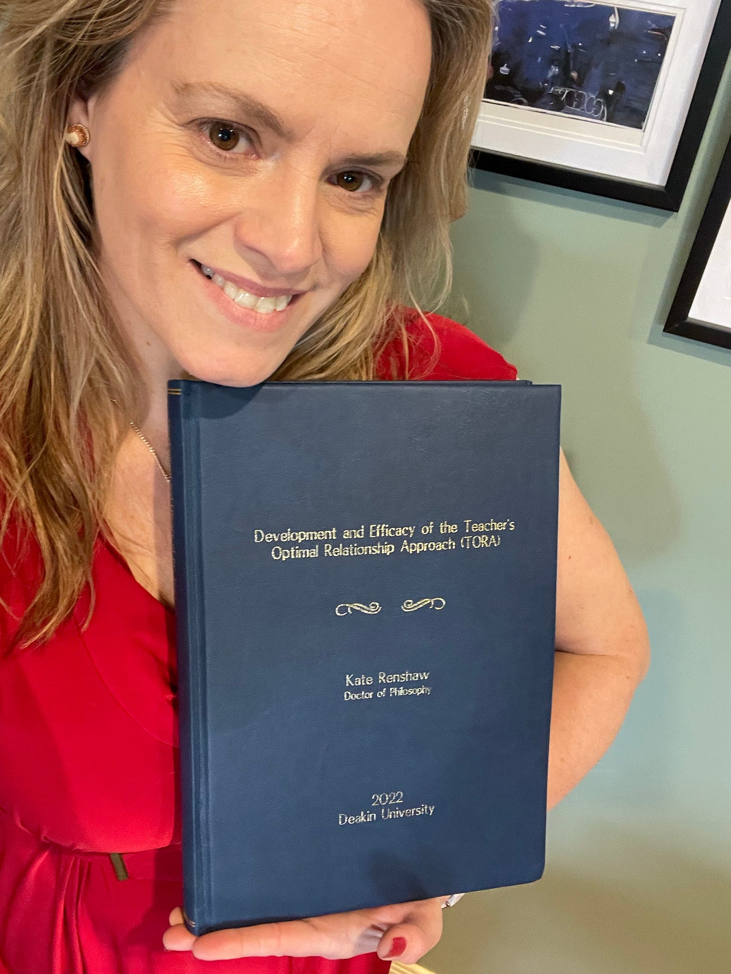 Dr Kate Renshaw smiling, holding her hardbound PhD thesis "Development and Efficacy of the Teacher's Optimal Relationship Approach (TORA)," Kate Renshaw, Doctor of Philosophy, 2022, Deakin University, navy hardbound with gold lettering. Dr Kate Renshaw smiling, holding her hardbound PhD thesis "Development and Efficacy of the Teacher's Optimal Relationship Approach (TORA)," Kate Renshaw, Doctor of Philosophy, 2022, Deakin University, navy hardbound with gold lettering.