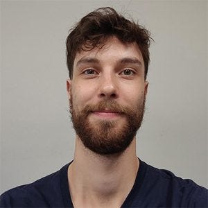 20260820_gabrielhenriquesavietto Um homem jovem com cabelo castanho ondulado e uma barba bem aparada, vestido com uma camiseta azul-marinho de gola em V, está sorrindo levemente e olhando para a câmera. Ele está posicionado no centro da imagem contra um fundo cinza claro uniforme