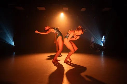 Deux danseuses en justaucorps sombres se produisent sur une scène plongée dans la pénombre. Sous un projecteur orange, l’une se cambre vers l’arrière, un bras tendu, tandis que l’autre se penche en avant, les bras repliés contre son corps. Leurs ombres s’allongent sur le sol. Deux danseuses en justaucorps sombres se produisent sur une scène plongée dans la pénombre. Sous un projecteur orange, l’une se cambre vers l’arrière, un bras tendu, tandis que l’autre se penche en avant, les bras repliés contre son corps. Leurs ombres s’allongent sur le sol.