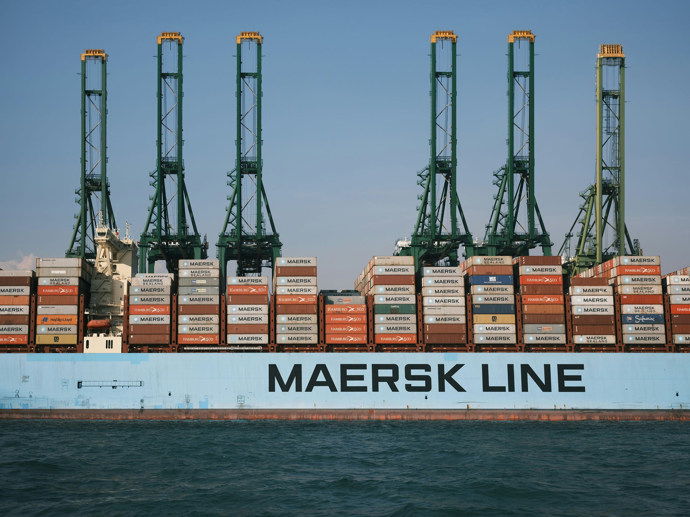 The photo of a large cargo ship is docked at a port, stacked high with colorful shipping containers. Several tall green cranes tower above the ship. The photo of a large cargo ship is docked at a port, stacked high with colorful shipping containers. Several tall green cranes tower above the ship.