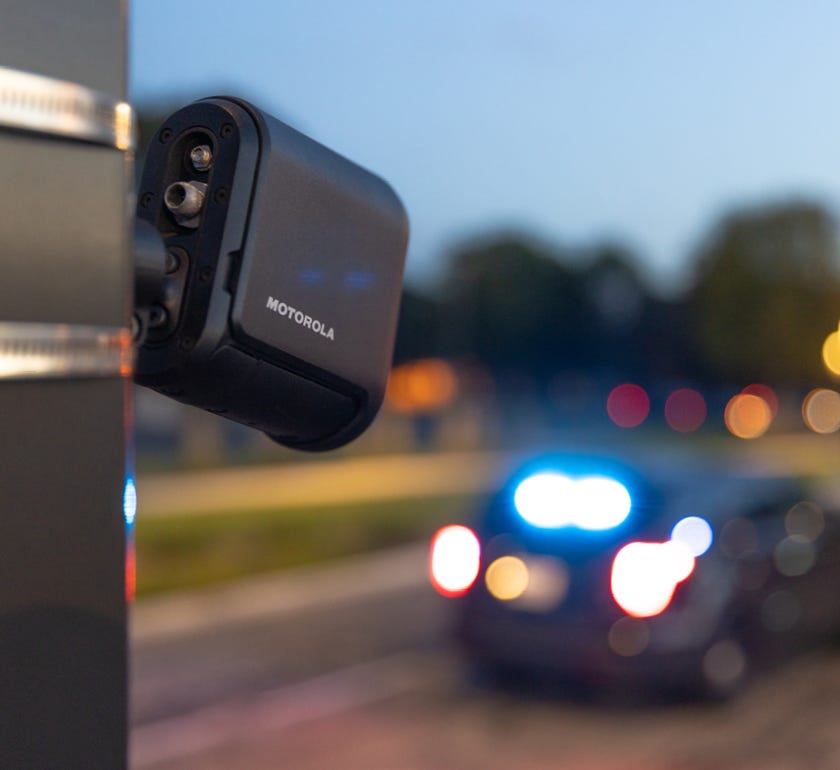 Motorola fixed-mounted license-plate reader attached to a pole, with a police vehicle and blurred roadway lights in the background.
