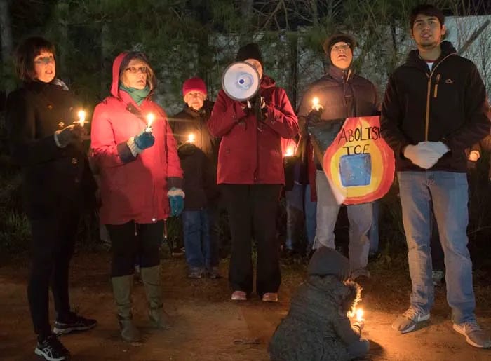 A group of people stand in candlelight, one person holds a megaphone and another with a sign that reads 'Abolish ICE' A group of people stand in candlelight, one person holds a megaphone and another with a sign that reads 'Abolish ICE'