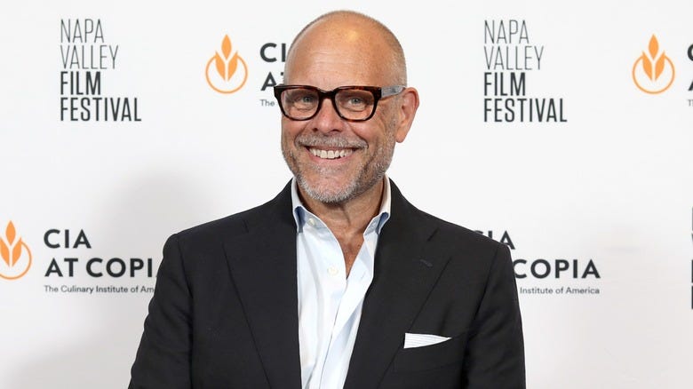 Alton Brown in interview chair on stage sitting above rug next to lamp.