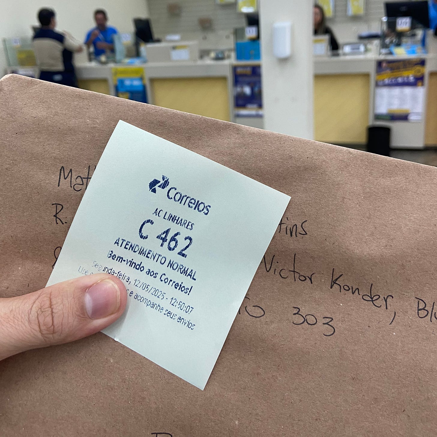 minha mão, dedo polegar de pele branca, segurando um envelope pardo e uma senha de atendido dos correios com número C462. ao fundo, os guichês de atendimento da agência dos correios e duas pessoas em desfoque.