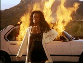 A woman in a white coat walks away calmly as a car behind her burns with large flames. Image source: Giphy