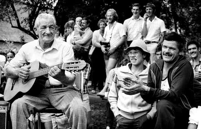 An elderly man plays the guitar while others stand and watch a performance we can't see. A man with missing front teeth claps his hands.