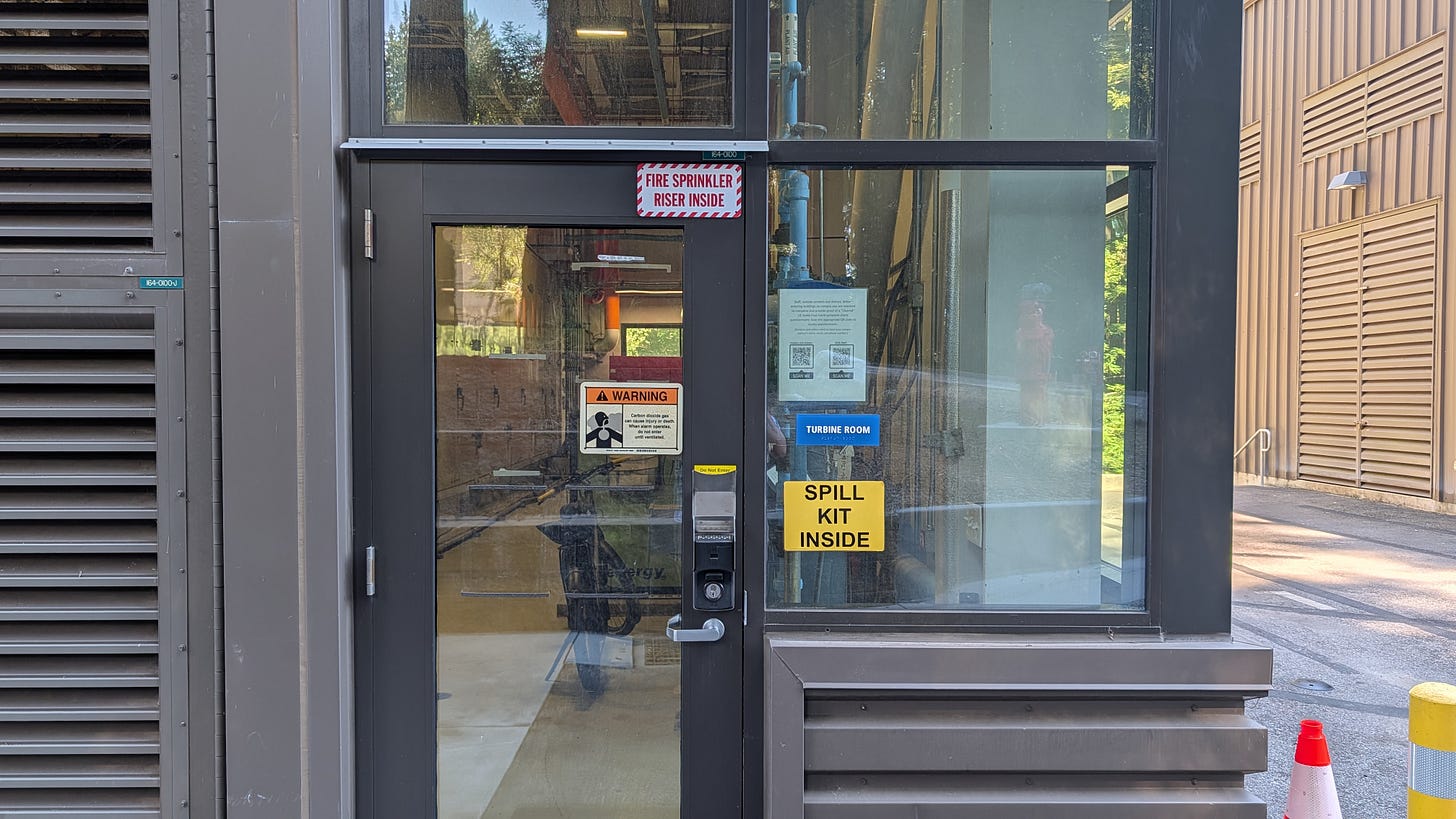 The glass entrance to the cogeneration plant's turbine room, with the machinery visible inside and warning signs mounted on the door and window.