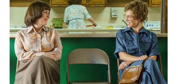 Two women in 1980s clothes sitting on folding metal chairs and smiling at each other