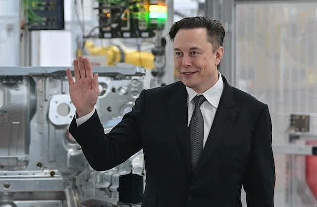 A white man in a suit holds up his right hand in front of car machinery A white man in a suit holds up his right hand in front of car machinery