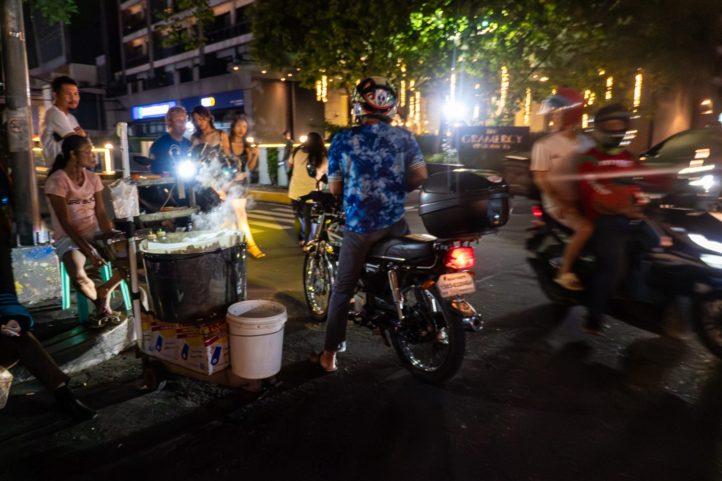 Busy streetcorner at night in Manila. Scooters are zooming by a vendor selling street food. A couple of scooter riders have stopped for food. The street vendor sits on a green plastic chair, wearing shorts, slippers, and a raggedy pink shirt. She looks tired. Drinks sit in a large metal tub set atop a cardboard box.