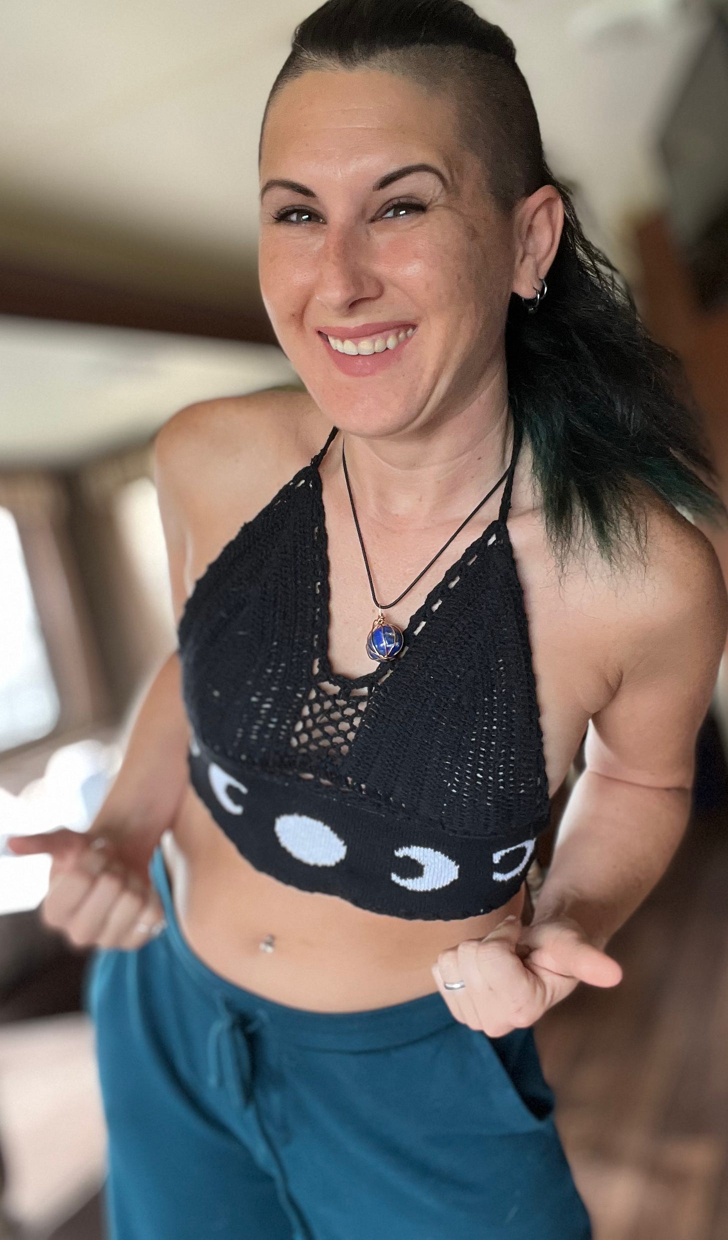 Happy Lyric, in teal pants, and a black top with moon cycles on it. They are in an RV, smiling and pointing at themselves.