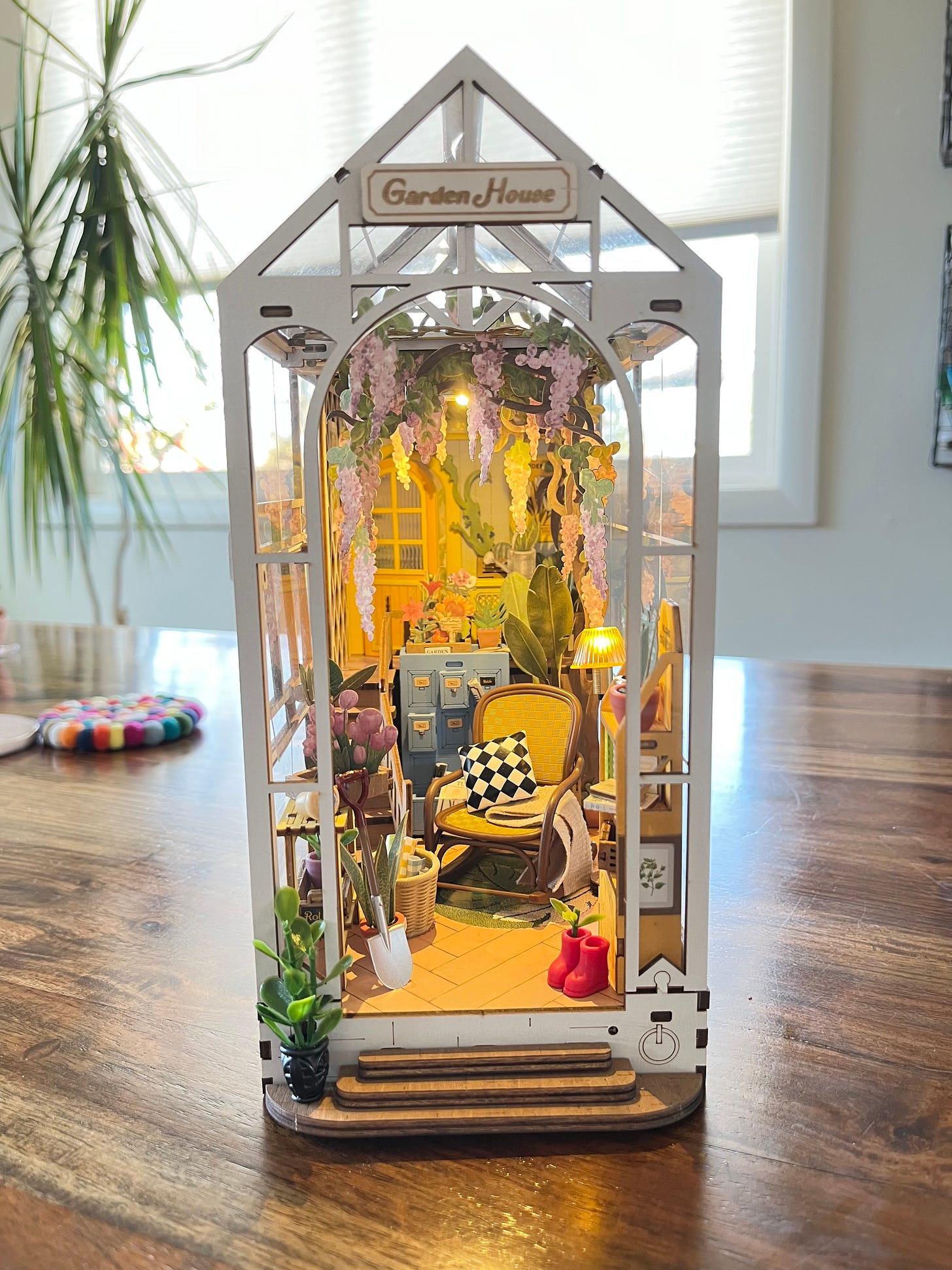 A photo of a miniature garden house complet with tiny lights that work. Paper lilacs hang down from the ceiling of a cozy greenhouse with plants, cupboards, and a rocking chair.