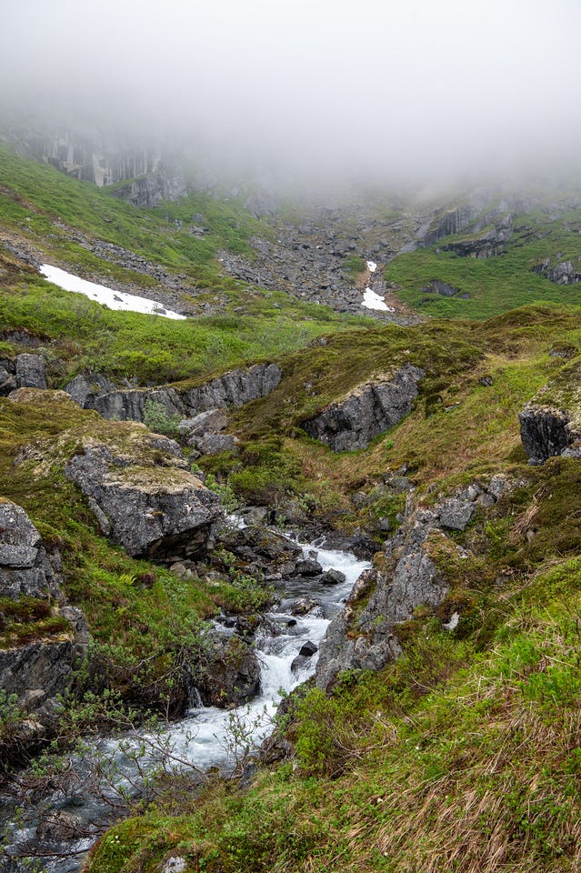 A whitewater creek tumbles down through a narrow rocky gully between banks of green tundra, brush, and grey outcrops. Above, the slope climbs past a patch of old snow toward a talus bowl, where thick fog swallows the upper mountainside, greying out the cliffs behind it. Everything is wet and saturated green.