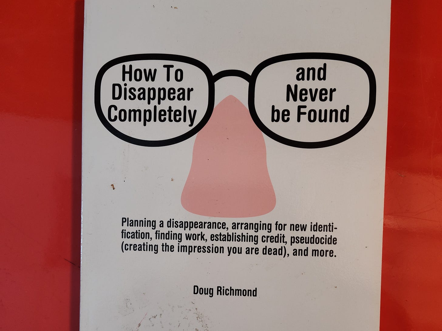 Book on orange table, title: How to Disappear Completely and Never Be Found: Planning a disappearance, arranging for new identification, finding work, establishing credit, pseudocide (creating the impression you are dead), and more.