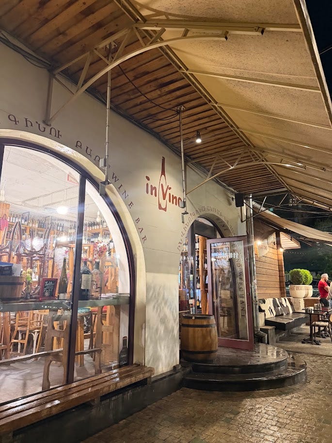 In Vino wine bar's arched storefront entrance at night in Yerevan, with warm interior lighting, wooden furniture, and a wine barrel by the door