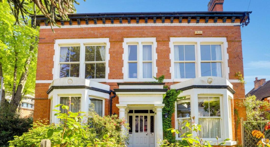 A large brick house with windows, a white door, and shrubbery next to it, with a blue sky above A large brick house with windows, a white door, and shrubbery next to it, with a blue sky above
