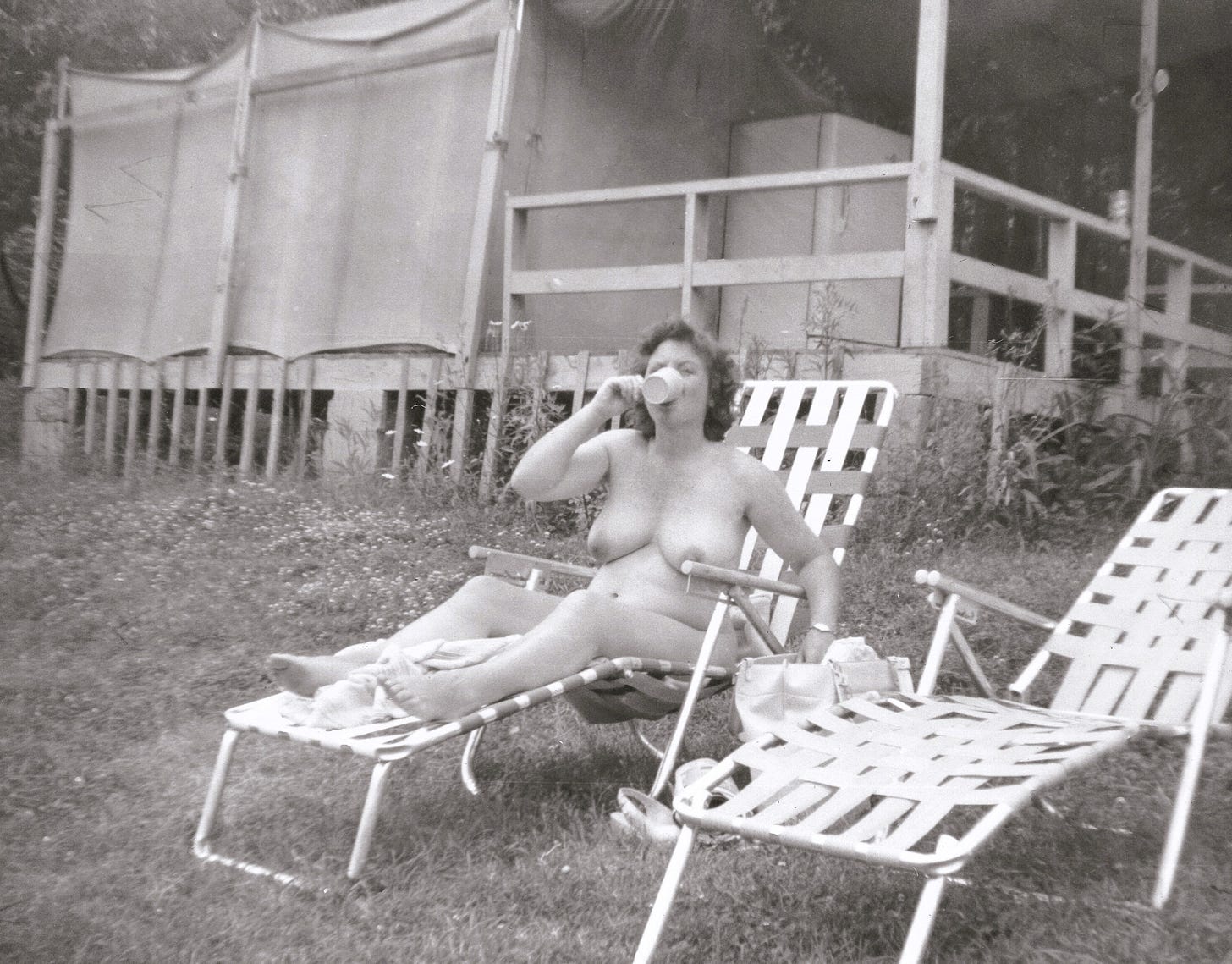 A nude woman lounges on a folding lawn chair outside a canvas-sided cabin, drinking from a cup.