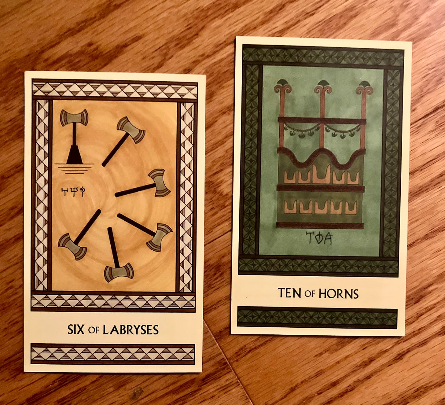 Two Minoan Tarot cards on a golden brown wood surface. The Six of Labryses is painted in shades of gold and cream. It shows five labryses flying through the air while a sixth labrys is in a stand, waiting for its turn to move. The Ten of Horns is painted in shades of green and brown. It shows a fancy garlanded platform where well-to-do people sit for public events.