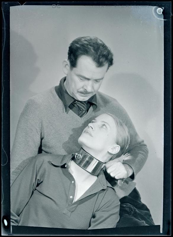 Black and white photo by Man Ray from his 1930 series “The Fantasies of Mr. Seabrook.” William Seabrook, a middle aged man in shirt and tie and sweater, stands over the photographer Lee Miller. Miller is a pretty young blonde woman, and she gazes up at Seabrook with a smile. Seabrook is holding a large metal restraining collar that is clamped around Miller’s neck. Black and white photo by Man Ray from his 1930 series “The Fantasies of Mr. Seabrook.” William Seabrook, a middle aged man in shirt and tie and sweater, stands over the photographer Lee Miller. Miller is a pretty young blonde woman, and she gazes up at Seabrook with a smile. Seabrook is holding a large metal restraining collar that is clamped around Miller’s neck.
