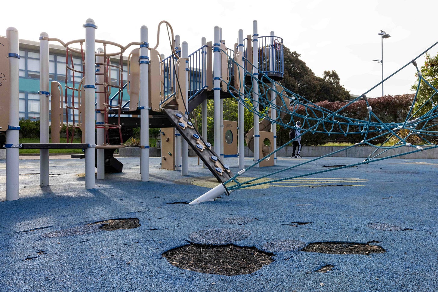 A playground with climbing equipment and rope structures sits on a blue surface with noticeable wear and holes. A person in the background walks nearby.