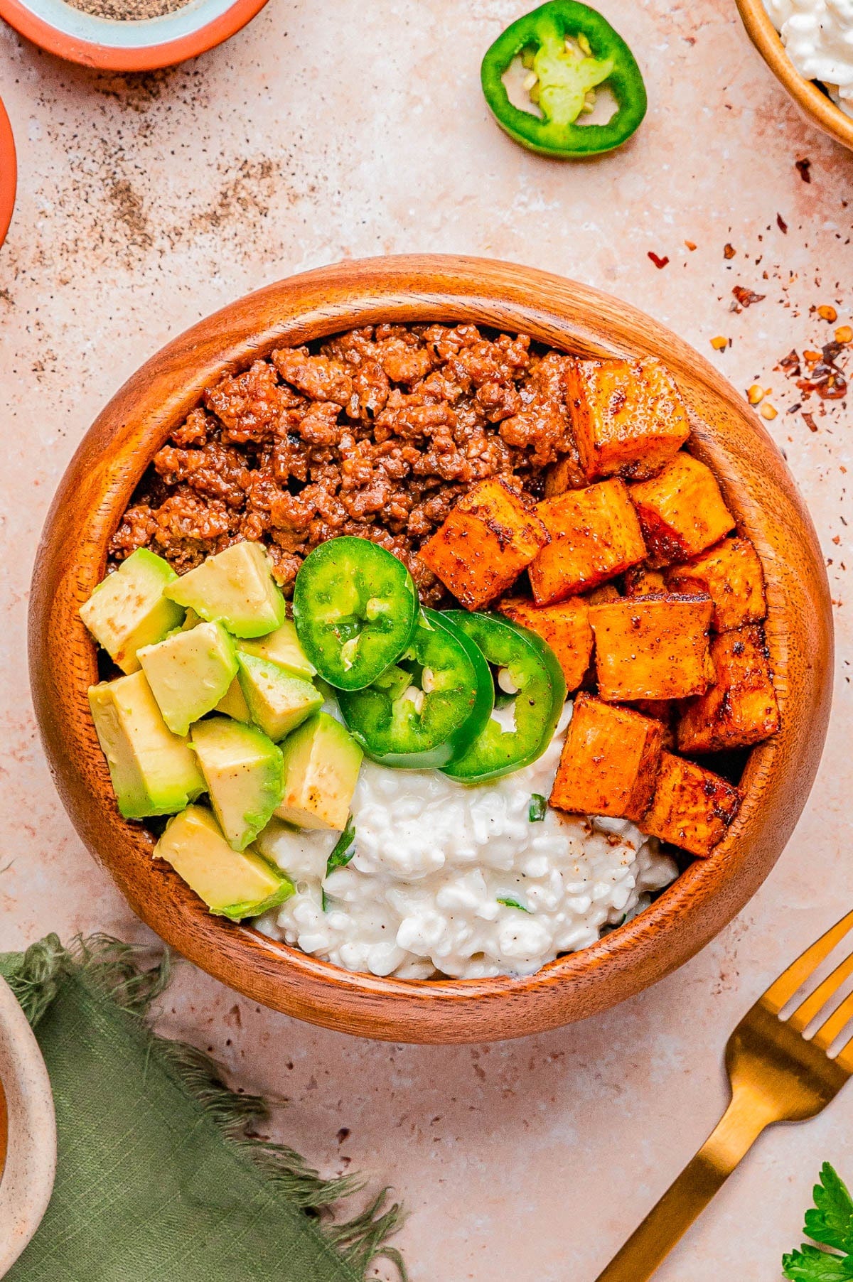 A wooden bowl containing seasoned ground meat, roasted sweet potatoes, diced avocado, cottage cheese, and sliced jalapeño, arranged in sections. A wooden bowl containing seasoned ground meat, roasted sweet potatoes, diced avocado, cottage cheese, and sliced jalapeño, arranged in sections.