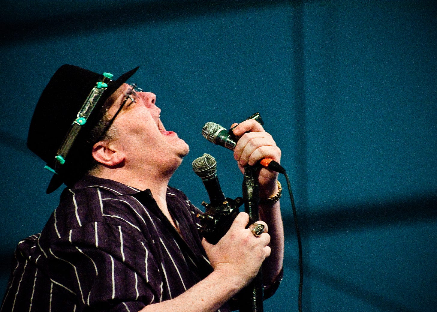 John Popper of Blues Traveler performs at the 2010 New Orleans Jazz & Heritage Festival, singing passionately into a microphone while holding his harmonica rig. He wears a black hat decorated with turquoise stones and a striped shirt. John Popper of Blues Traveler performs at the 2010 New Orleans Jazz & Heritage Festival, singing passionately into a microphone while holding his harmonica rig. He wears a black hat decorated with turquoise stones and a striped shirt.