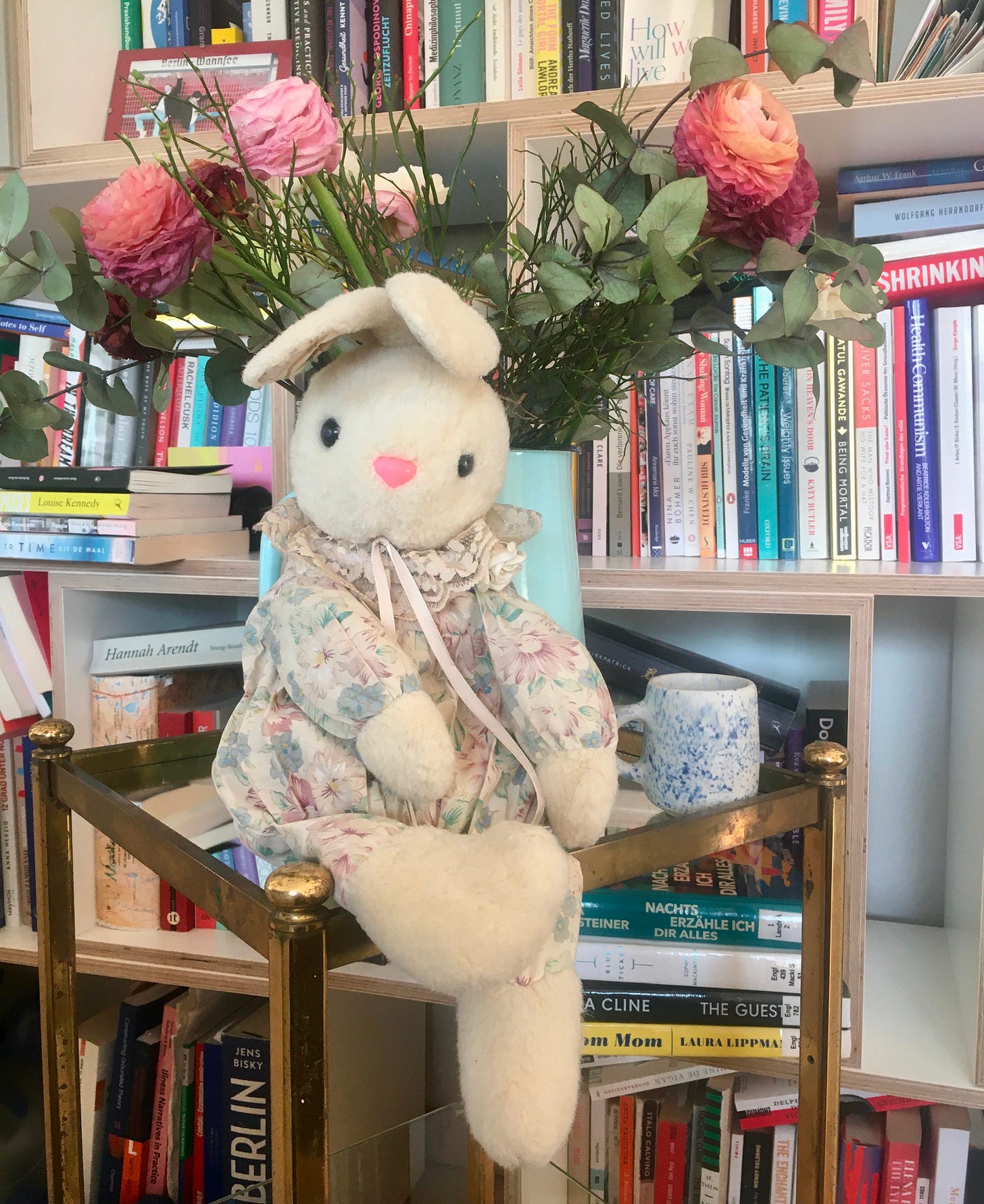 Ein Kuschelhase sitzt auf einem kleinen Tisch vor einer Blumenvase, daneben steht eine Tasse. Im Hindergrund ein sehr volles Bücherregal.
