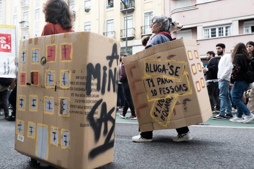 Una fotografia scattata durante una manifestazione in strada a Lisbona. Due manifestanti camminano indossando grandi scatole di cartone trasformate in costumi che rappresentano condomini, con finestre disegnate a mano. Sulla scatola di destra sono incollati cartelli gialli con scritte di protesta in portoghese, tra cui 'ALUGA-SE T1 PARA 10 PESSOAS' (Affittasi T1 per 10 persone) e '€1.100 P/ QUARTO' (1.100€ per stanza), a denunciare il caro affitti.