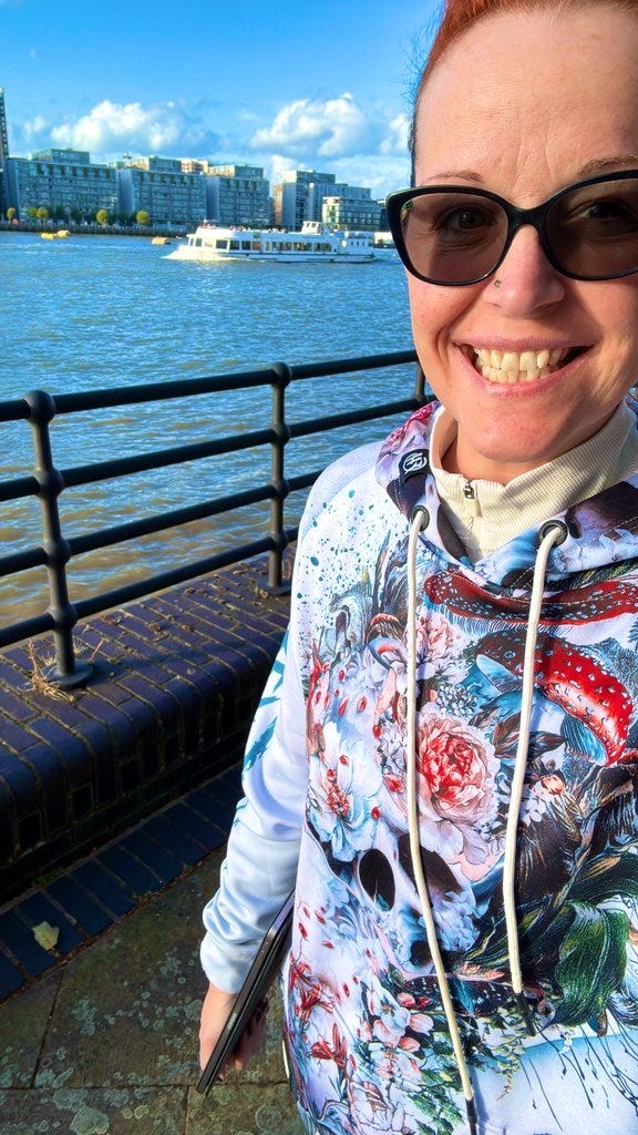 A smiling selfie of Georgina Dean walking along the Thames in London, wearing a colourful hoodie. The water and city buildings are visible behind her, conveying a sense of thoughtful, active reflection.