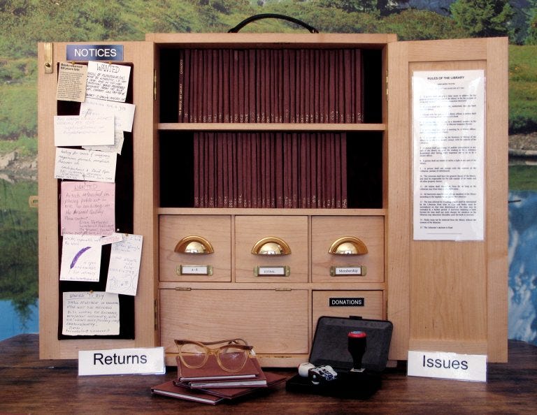 A small wooden cabinet, shown open. In the center are two shelves of uniform books, all with narrow, brown spines. Beneath the shelves is a card catalogue. On the inner doors are posted notices.