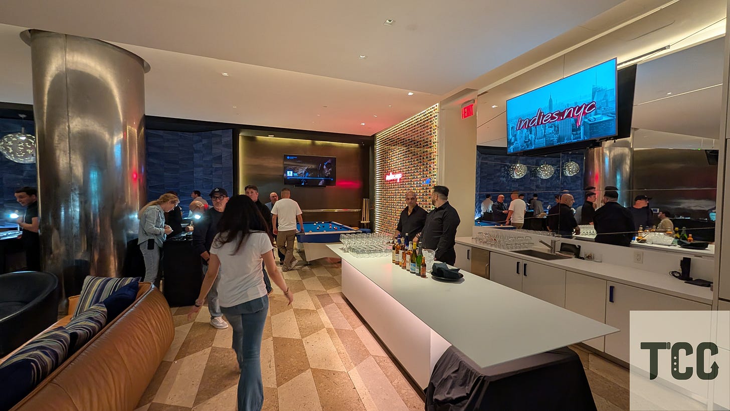Interior shot of the Indies event space featuring a sleek modern bar, guests mingling around cocktail tables, and a screen above the bar displaying the “indies.nyc” logo over a cityscape. The vibe is upscale and social.
