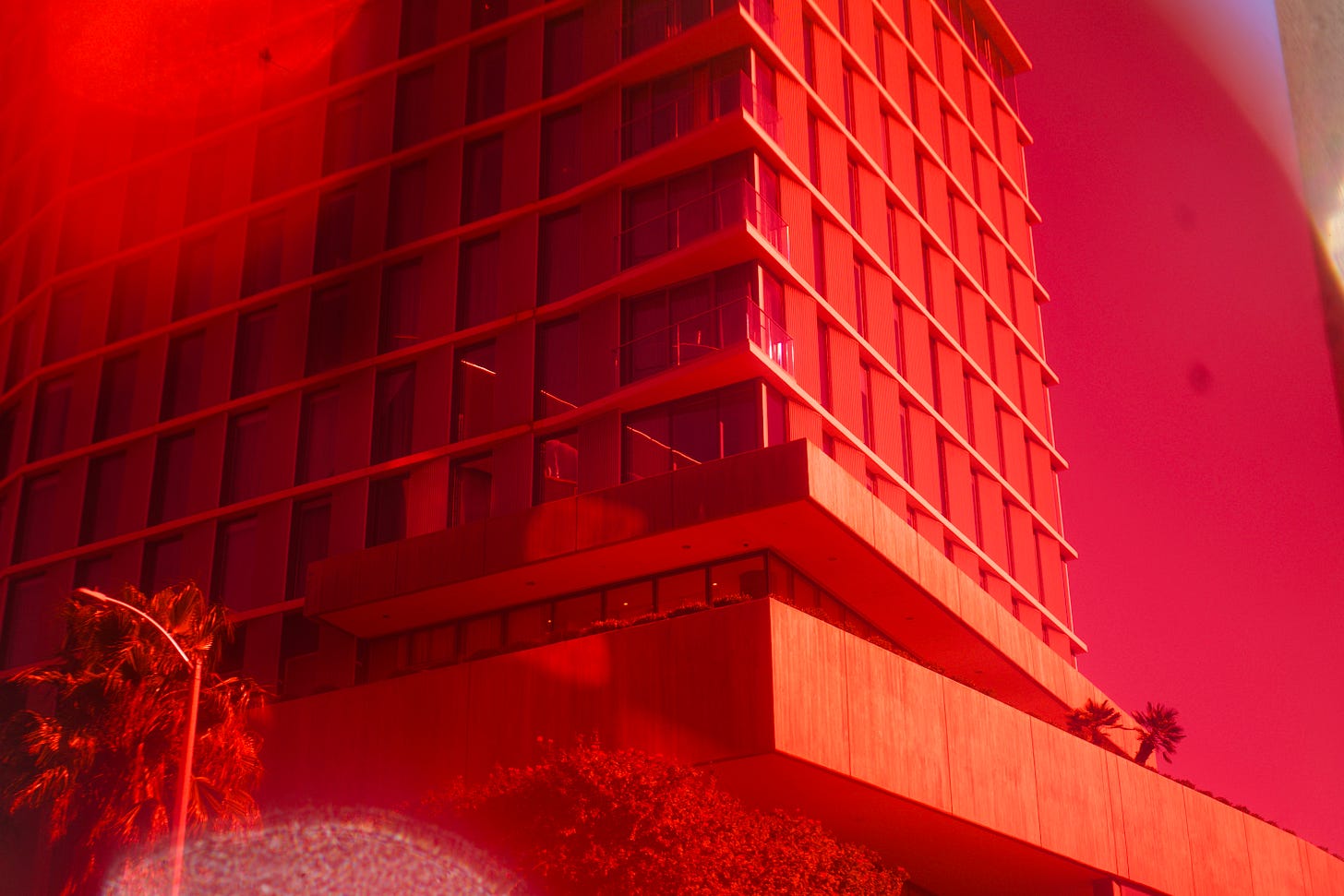 Modern high-rise building viewed from below, washed in a deep red glow with palm trees at the base and geometric lines rising into the sky.