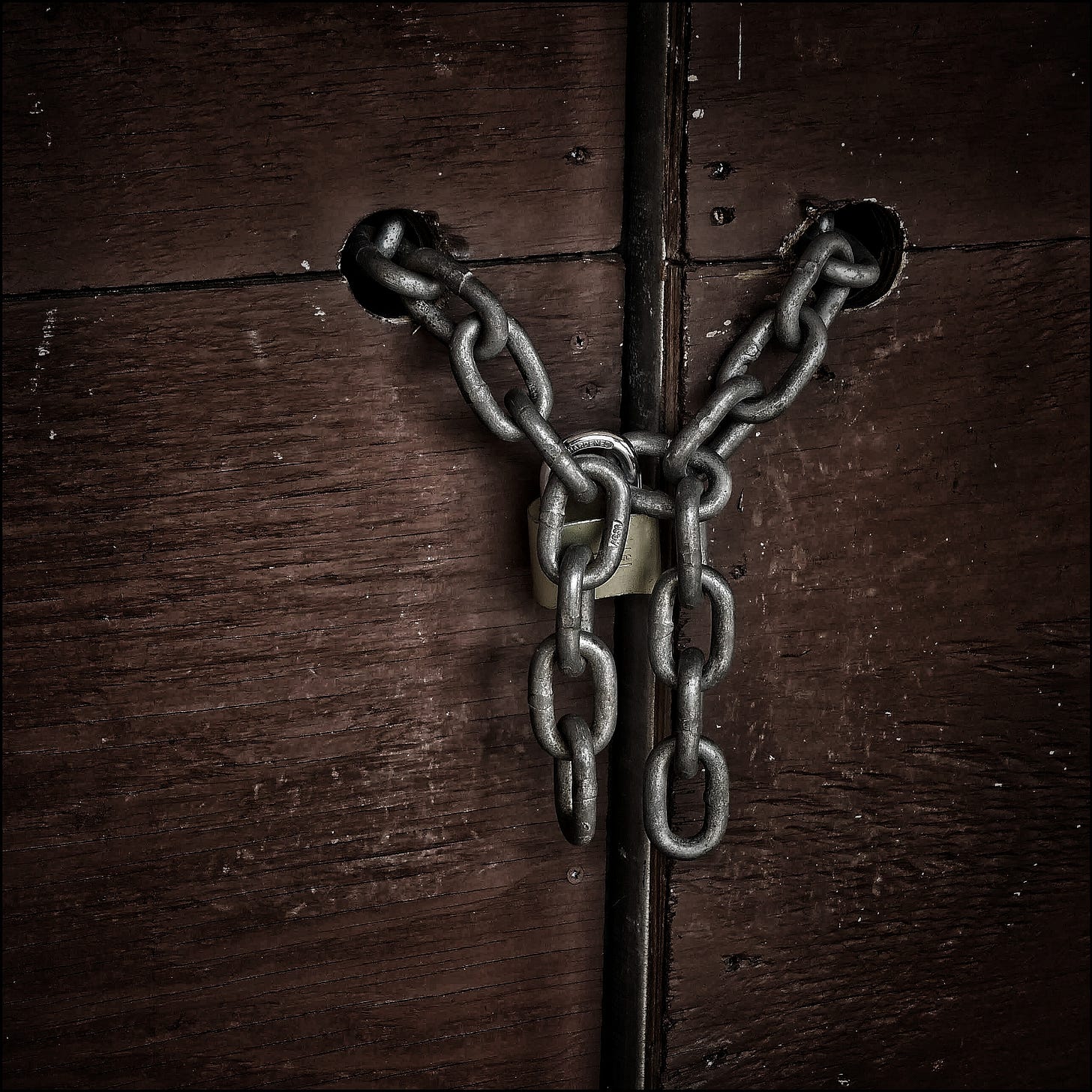 A photo of an old, worn wooden gate, or perhaps the door of a barn. It is secured with a strong chain, for some reason…