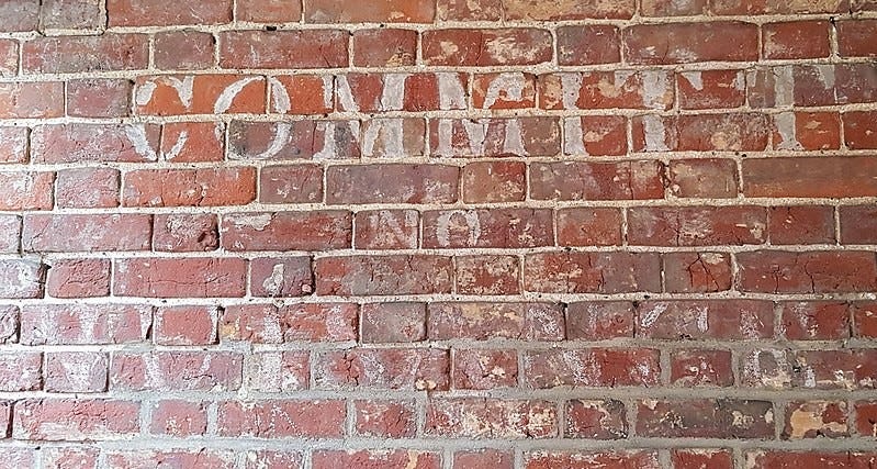 A brick wall with a very faded slogan in white paint which reads “Committ [sic] no nuisance”