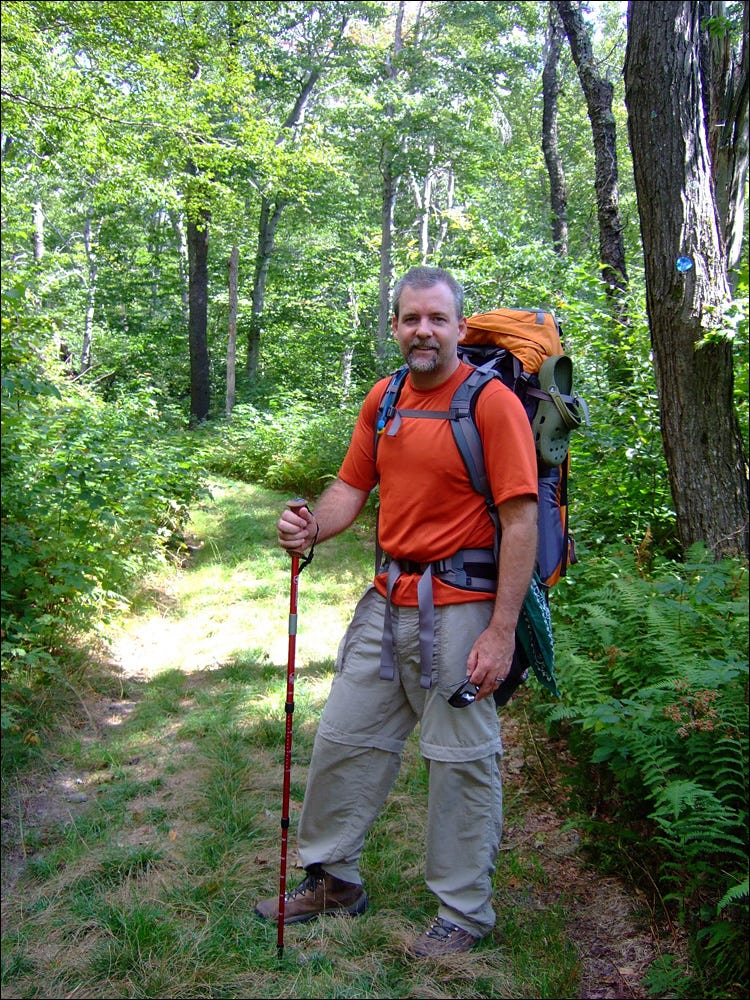 The author stands on grassy Spruceton Trail, leafy green trees all around