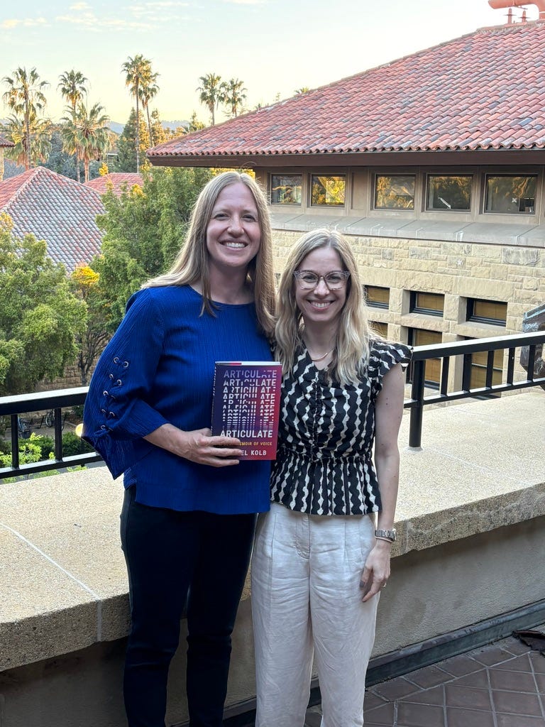 Lindsey Dolich Felt and I stand on a terrace at Stanford, the Quad and several palm trees visible in the background. We are smiling and holding a copy of my book ARTICULATE. I'm in a deep blue shirt, and she's in a patterned black-and-white blouse.