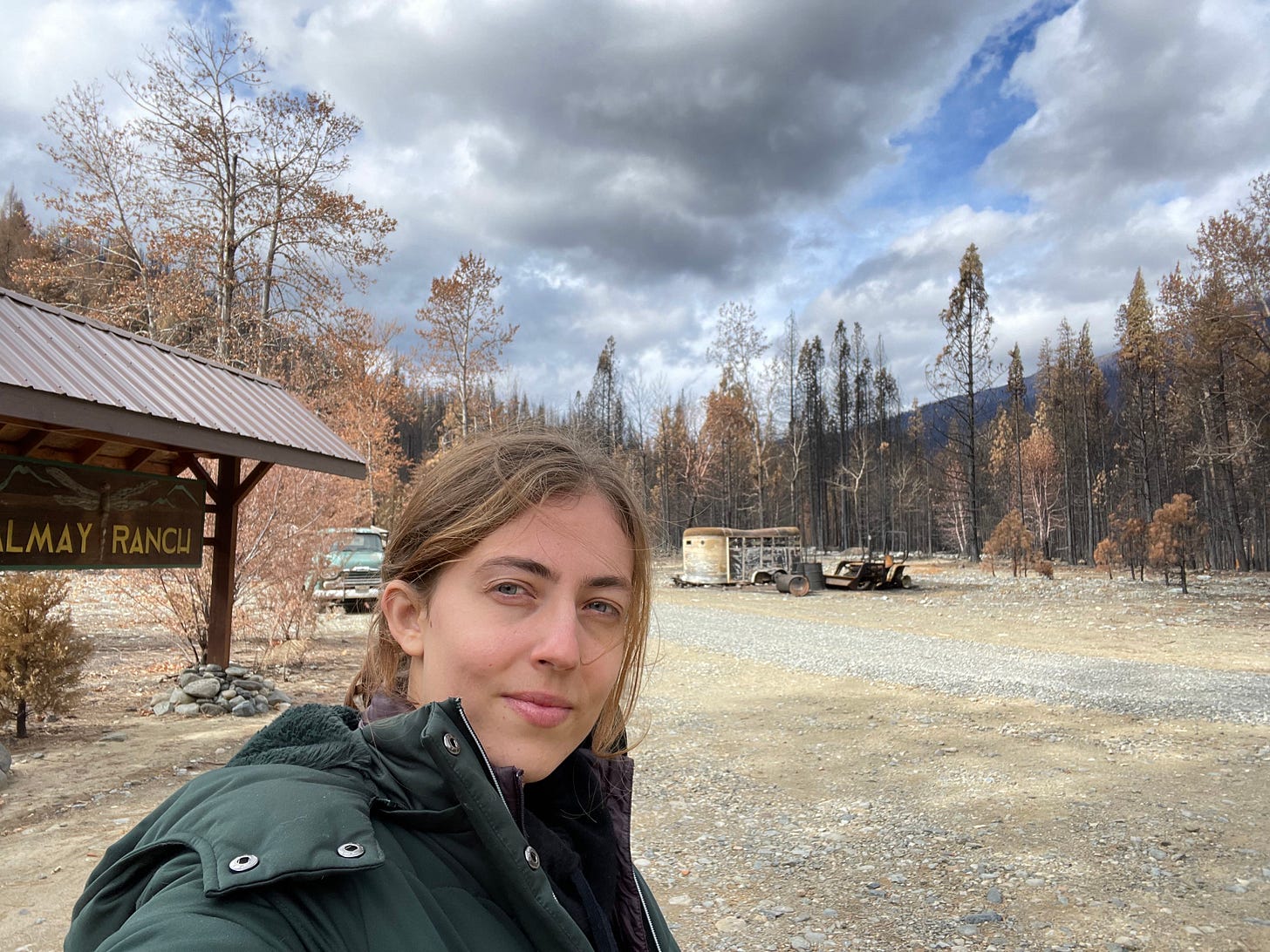 Ana, a blonde Caucasian woman, takes a selfie in front of an open space in the aftermath of a fire. There is a burnt-out car in the background.