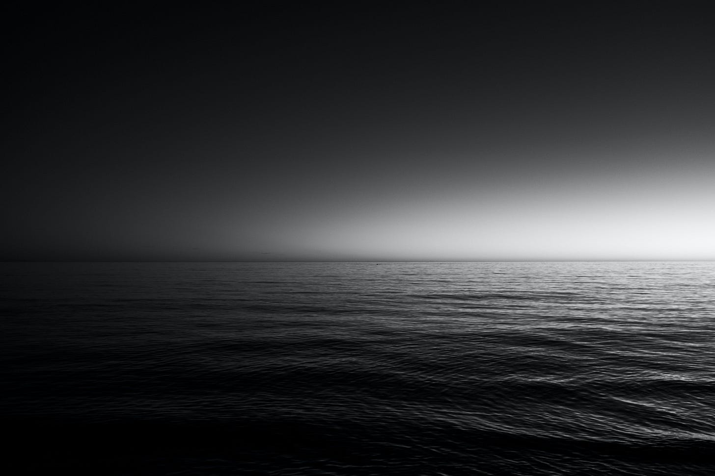 A black and white photo of a body of water with gentle waves rippling over the top. The sky is dark with a little bit of light on the horizon. The water is also dark. A black and white photo of a body of water with gentle waves rippling over the top. The sky is dark with a little bit of light on the horizon. The water is also dark.