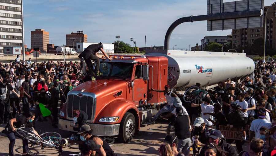 A red semi-truck is stopped and surrounded by a large protesting group on the I-35W bridge over the Mississippi River. It is a chaotic scene with a protestor climbing on the hood of the truck and others shoving bikes into the undercarriage.