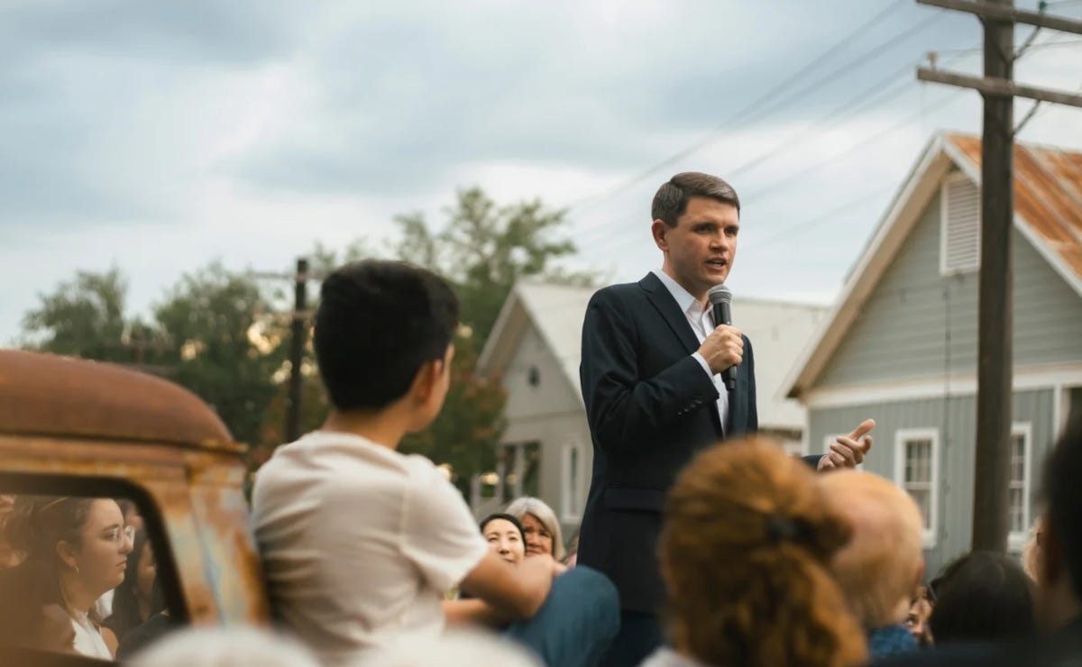 Man standing on the back of a pickup truck, holding a microphoone and speaking to a crowd