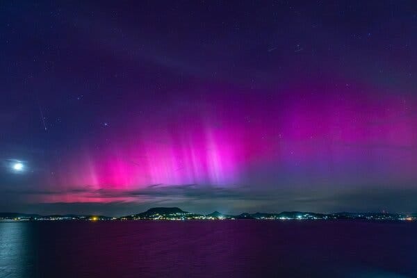 Vibrant pink and purple colors streak across the sky above a lake with city lights and mountains on the shoreline.