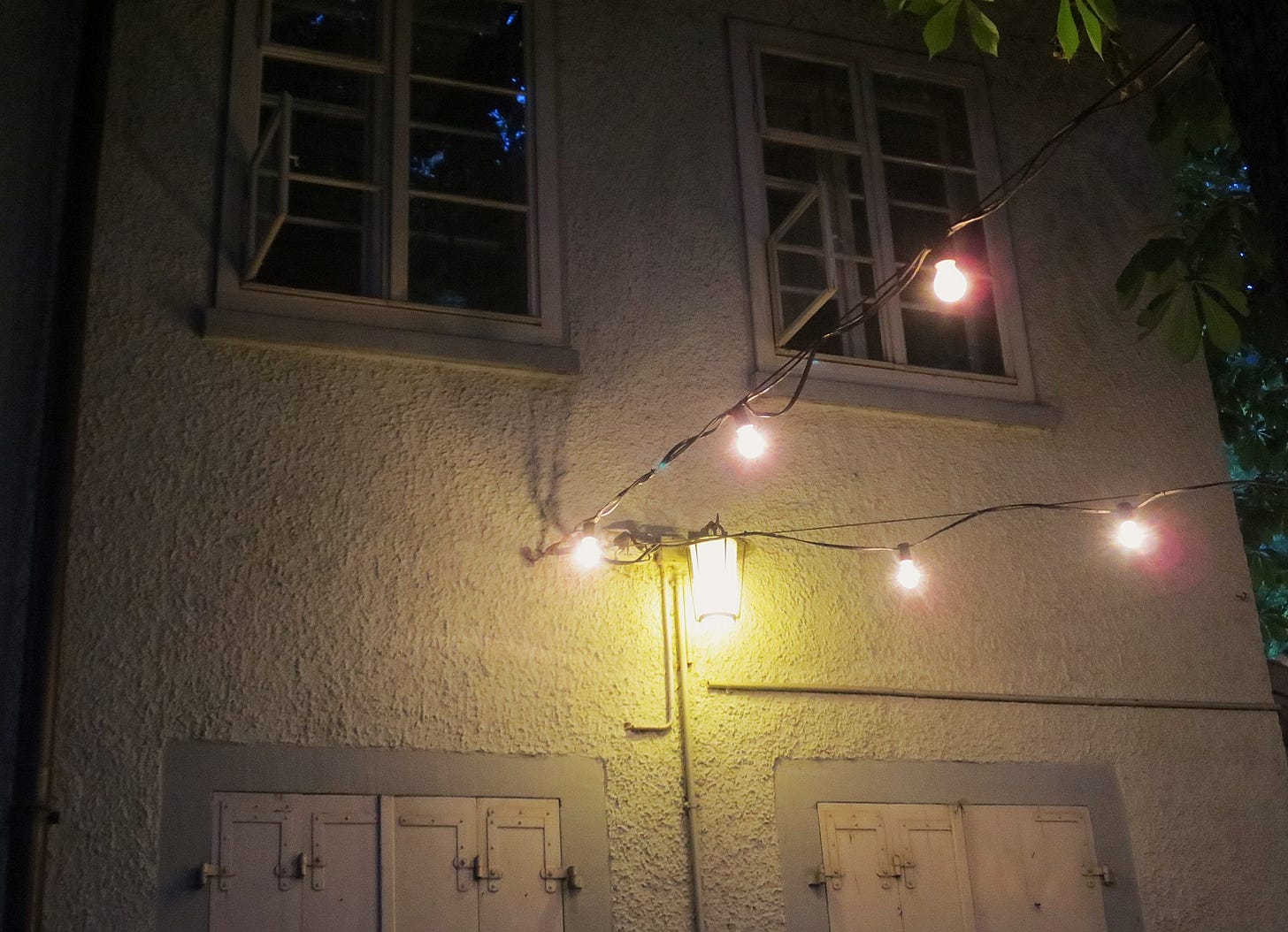 Fotografia notturna dove le luci elettriche sono appese a dei fili fissati ai rami degli alberi e al muro bianco di un vecchio edificio, di cui si vedono anche alcune finestre con le persiane bianche di legno