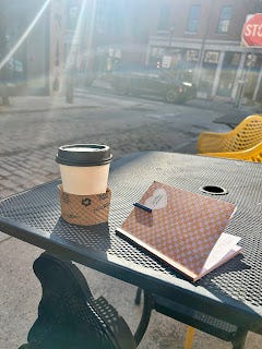 A sunny cafe table with a paper cup and a notebook.  My foot, in a black boot, is underneath.
